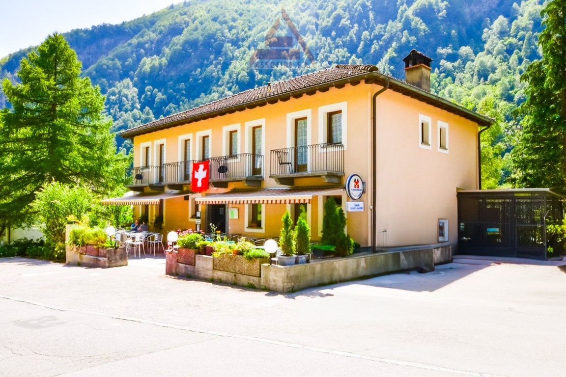 2-story building with a yellow exterior, balconies, and a Swiss flag. The building is surrounded by trees and has a patio area in front with tables and chairs. The building appears to be a hotel or restaurant located in a mountainous, forested area.