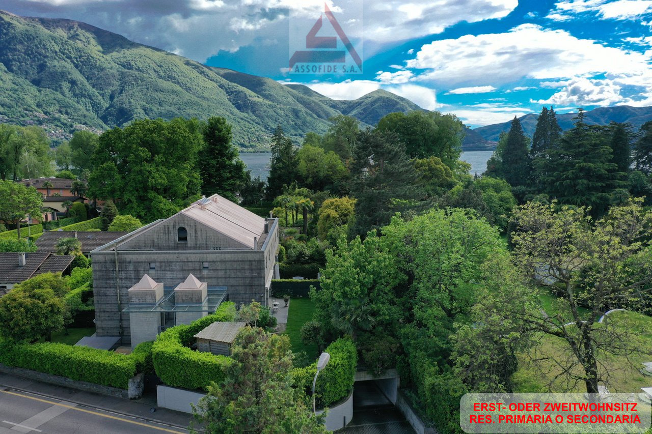 A two-story villa with a view of the mountains and lake, large landscaped garden, and a nearby road.