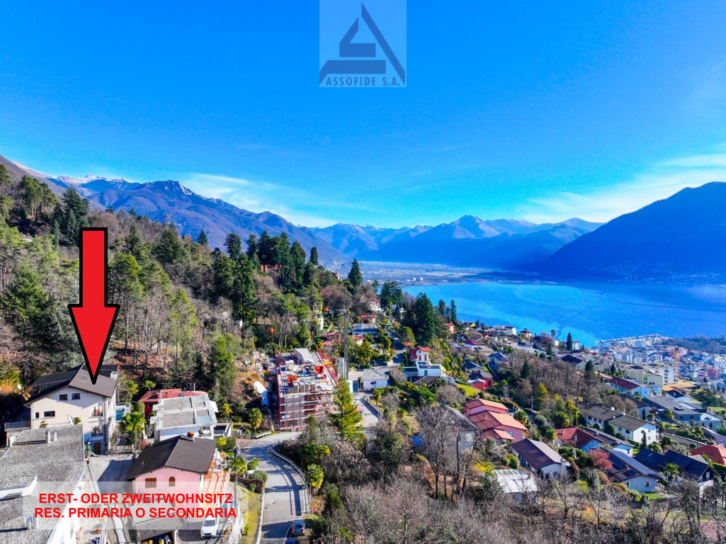 A hillside residential area with buildings and trees, a lake and mountains in the background, a red arrow pointing to a house on the left.