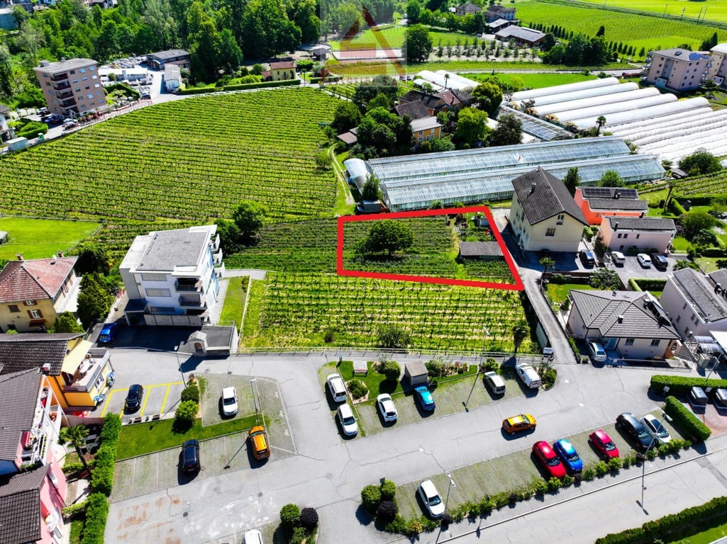 The image shows an aerial view of a residential area surrounded by vineyards and greenery. There is a parking lot visible in the foreground, and several buildings and houses can be seen in the background. The area appears to be a small town or village nes