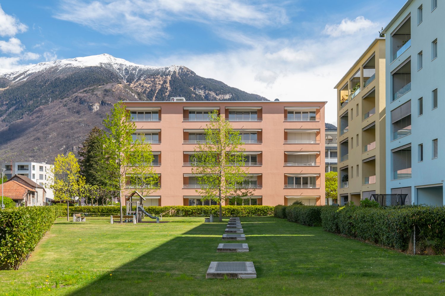Modern apartment building with multiple floors, surrounded by greenery and a playground, with a view of the mountains