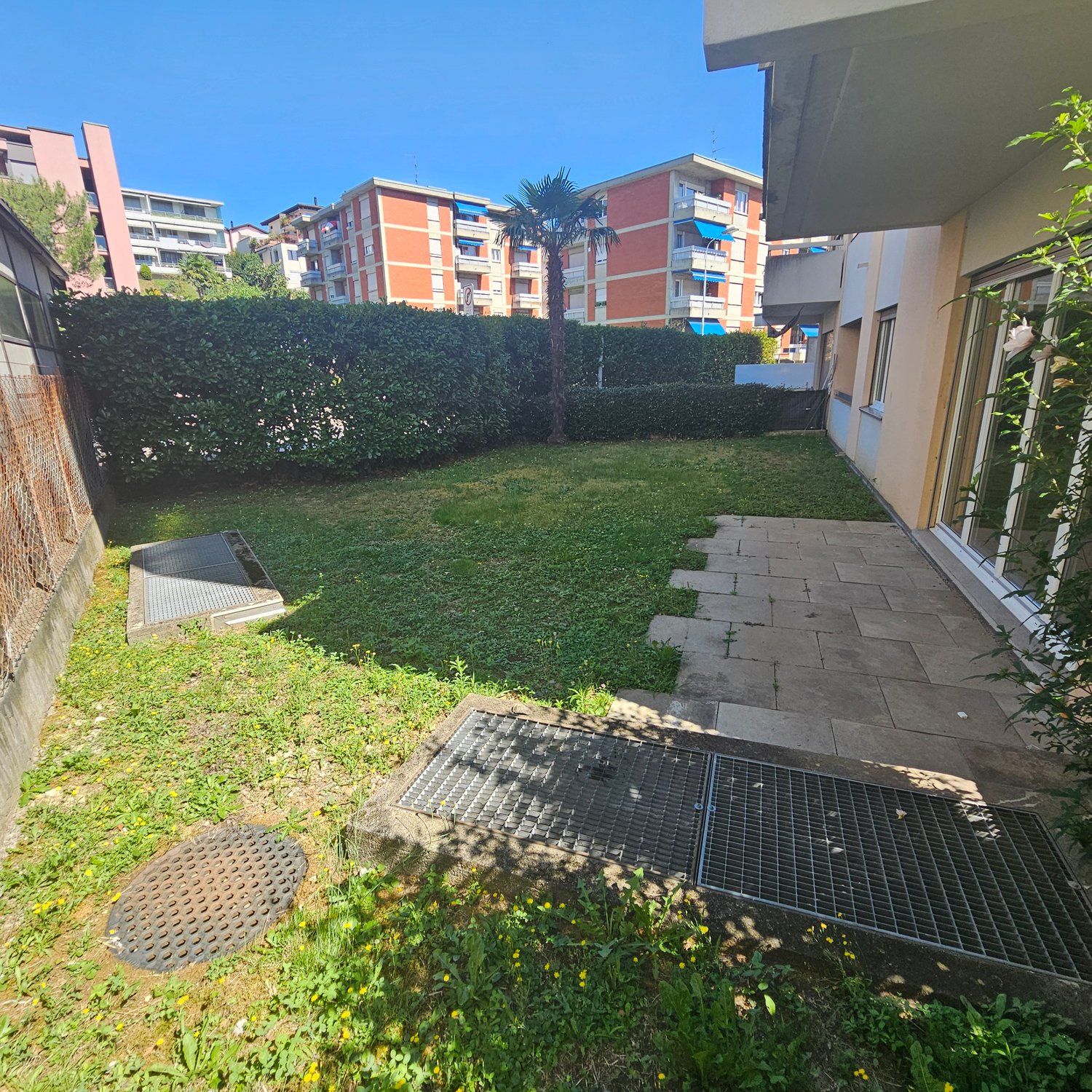 The image shows a residential apartment complex with several multi-story buildings. The buildings have a mix of orange and gray colors. There is a small grassy area with a palm tree in the foreground, and a paved walkway leading to the buildings. The comp