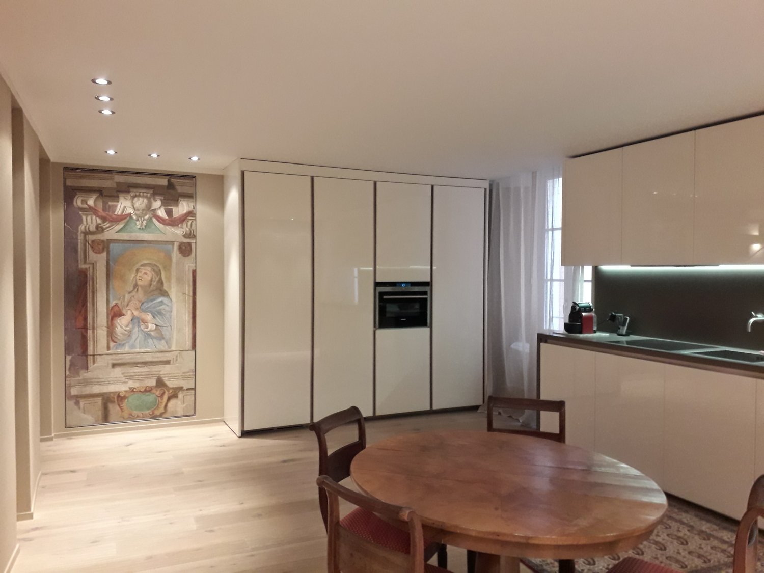 white cabinets, wooden table, chairs, integrated oven, large artwork, modern lighting