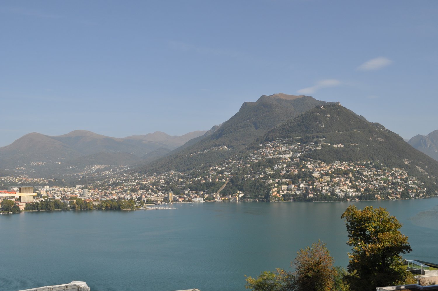 Scenic view of a mountainous town situated on the shores of a large lake, with blue waters and a clear sky in the background.