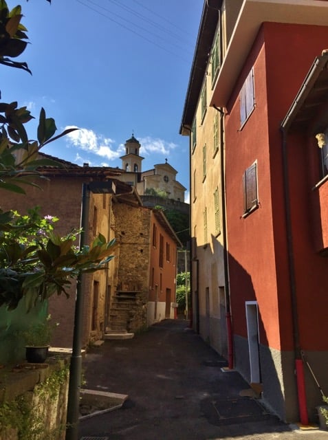 Narrow alleyway with colorful buildings, stone walls, and a church steeple visible in the background. The scene has a charming, historic European feel with lush greenery and a bright, sunny sky.
