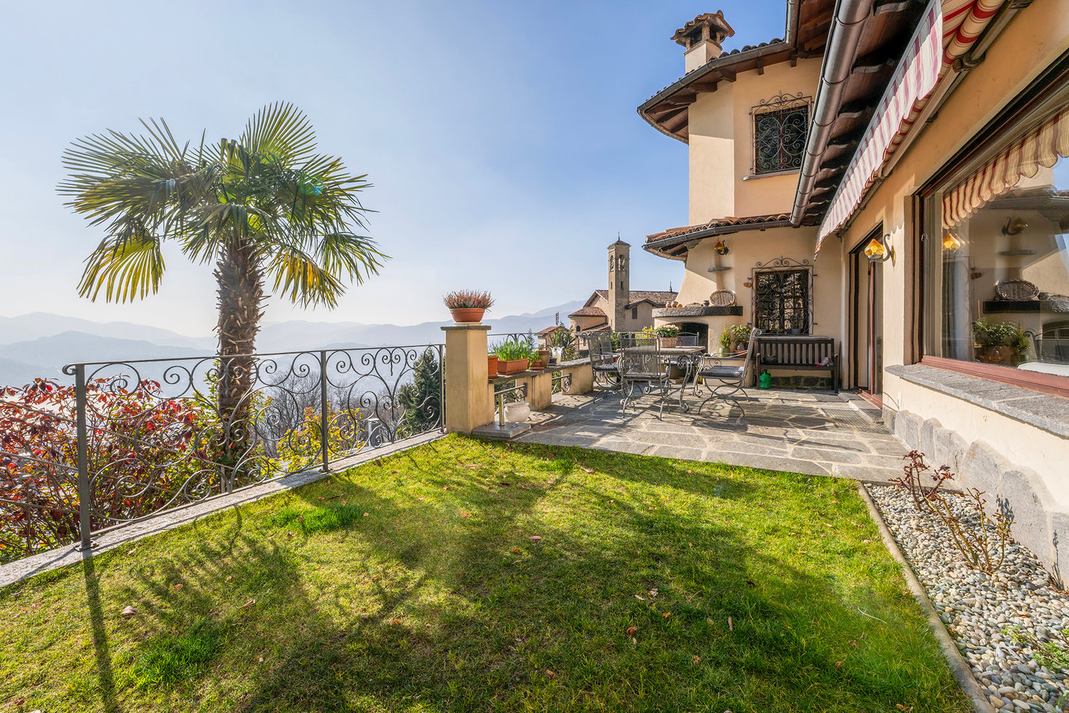 Two-story Mediterranean-style house with a terrace, palm tree, and ornate wrought-iron railing overlooking a grassy yard and mountains in the background. The house has a stone exterior, red tile roof, and decorative architectural details.