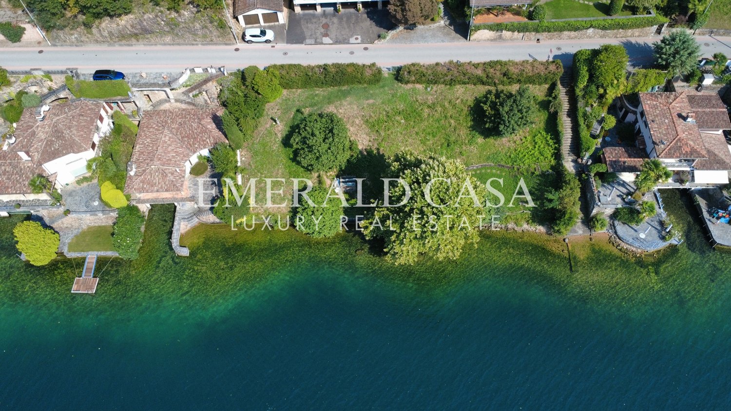 Aerial view of a lake with a villa property on the right, large swimming pool, wooden deck, surrounded by greenery and trees, and a lake in front.