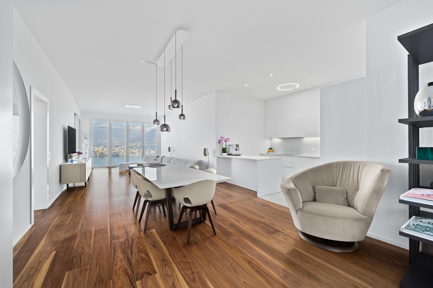 modern interior, hardwood floor, large windows, dining table with chairs, white walls, white cabinets, built-in television, floral vase, modern chandeliers, contemporary armchair, black shelf