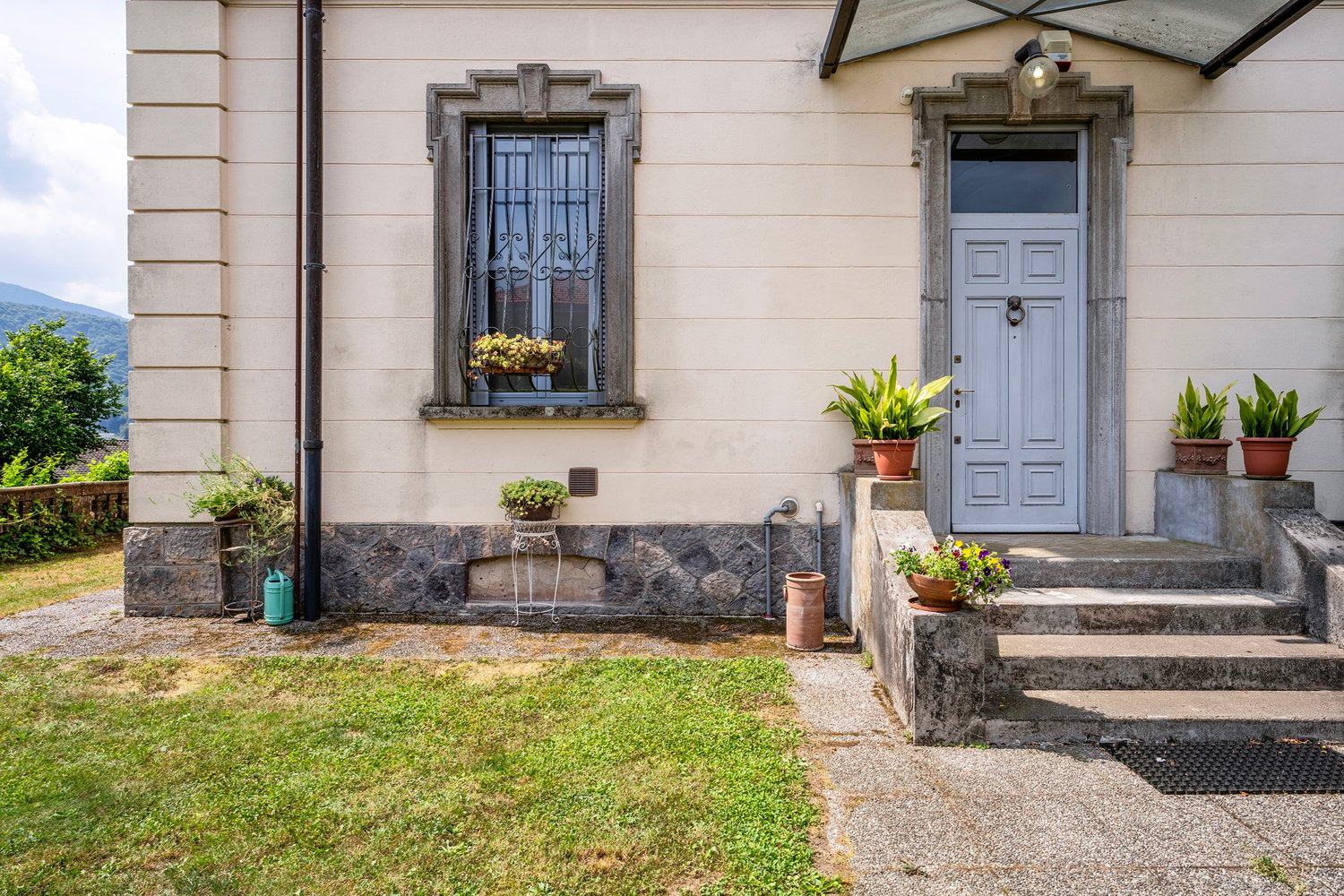 Traditional house, cream painted exterior, stone wall base, front door with window, step, several plants and flowers in front