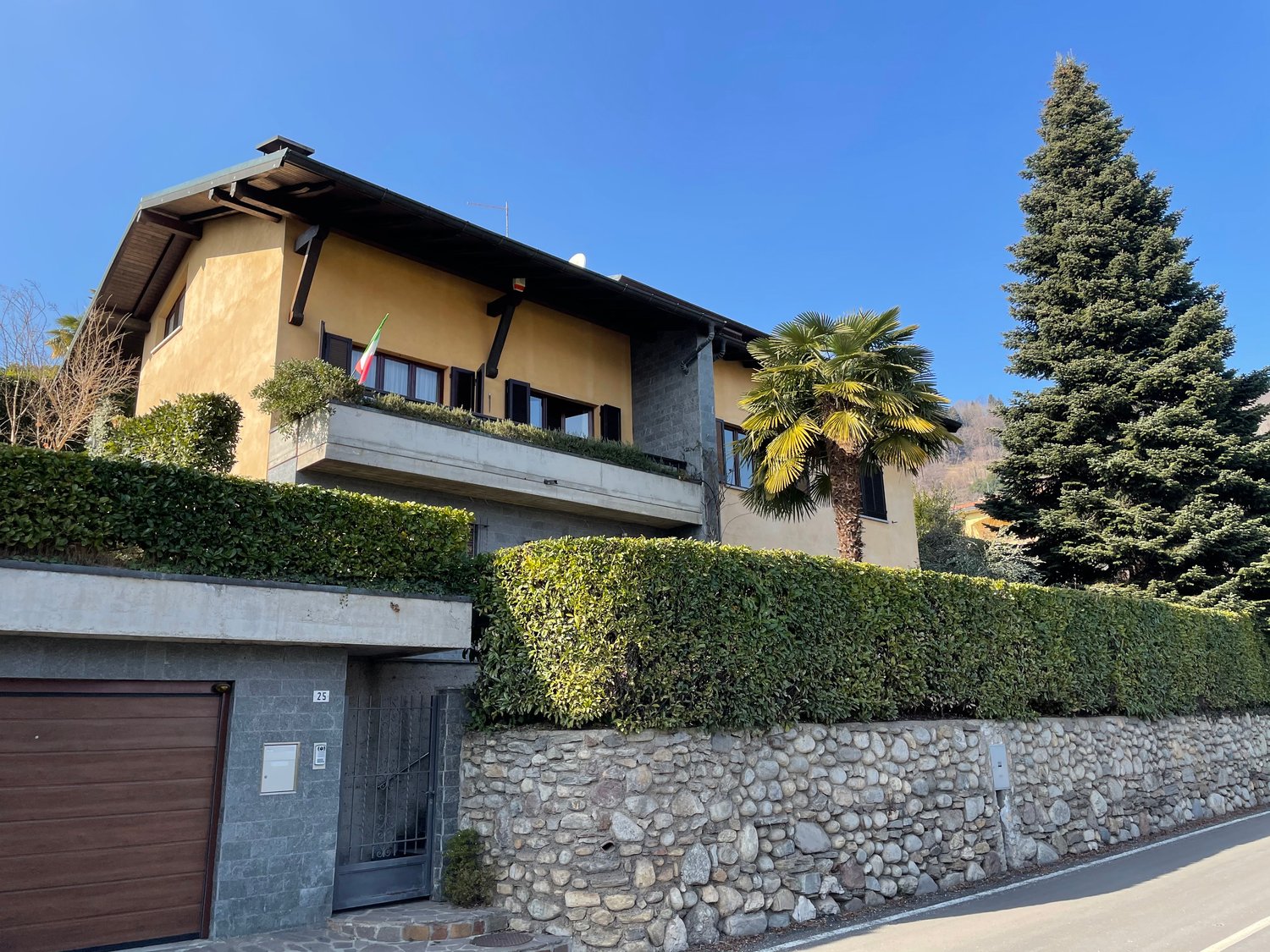 Detached house, beige painted exterior, small balcony, large garage, landscaped hedges, mature trees