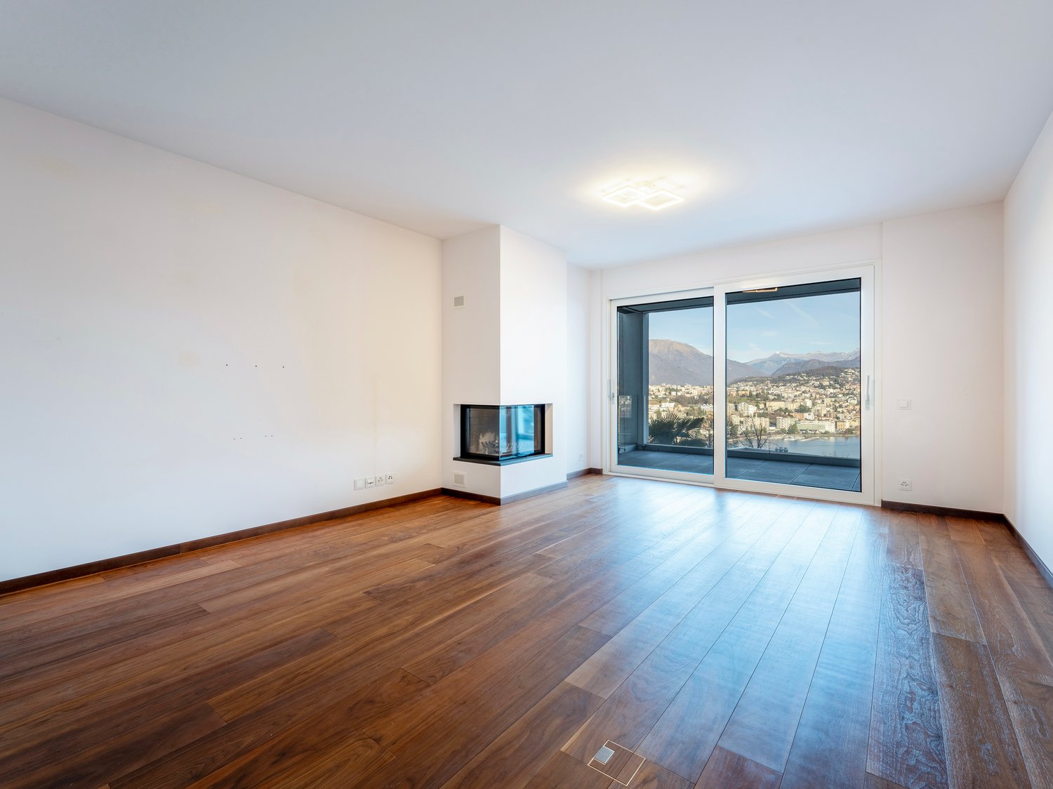 Empty room with hardwood floors, white walls, fireplace, sliding glass doors to balcony, city view.