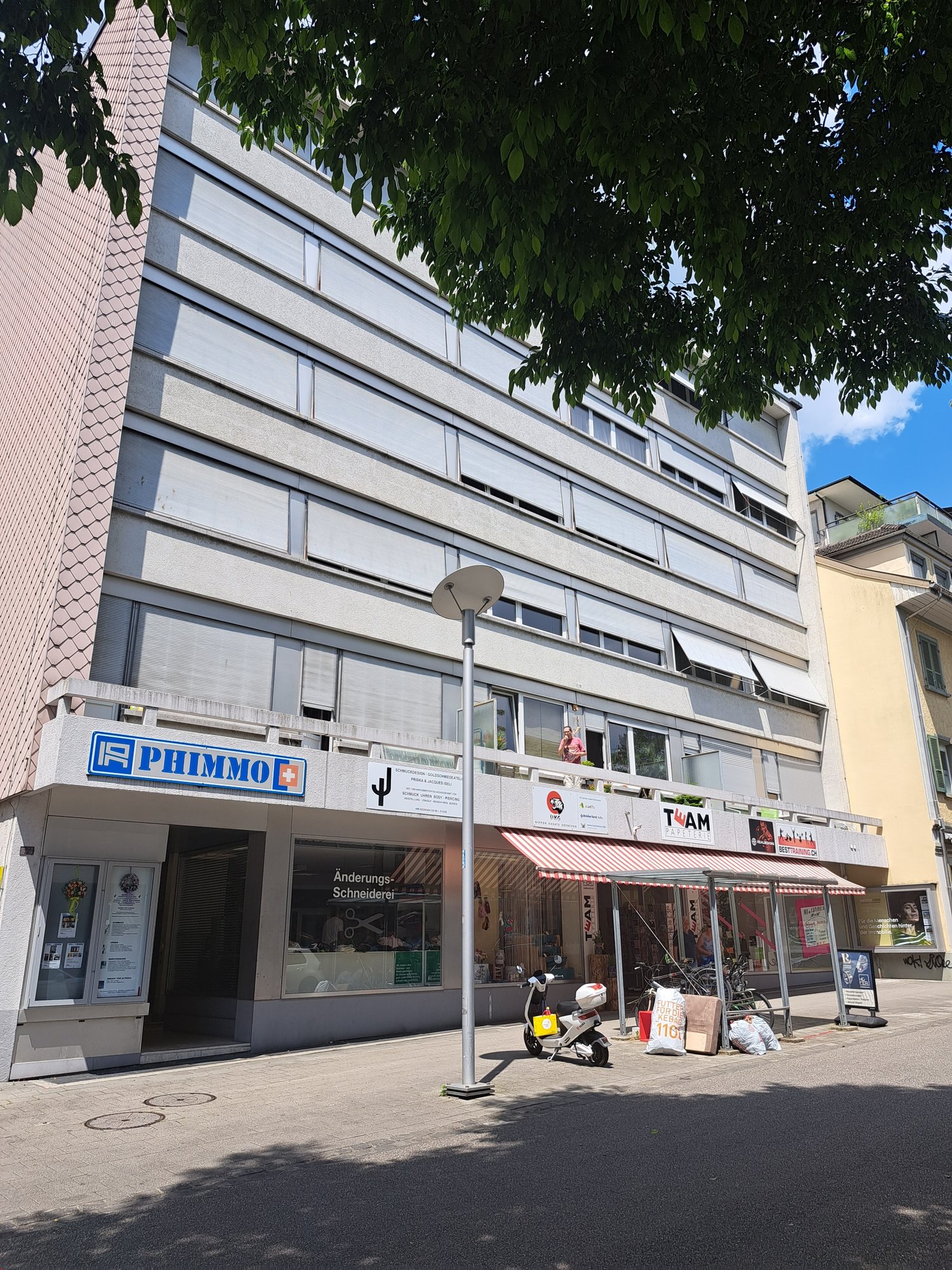 concrete and glass building, multiple windows, sign reads Phimmo, awnings, store front, lamp post, scooters, trash cans, bicycles
