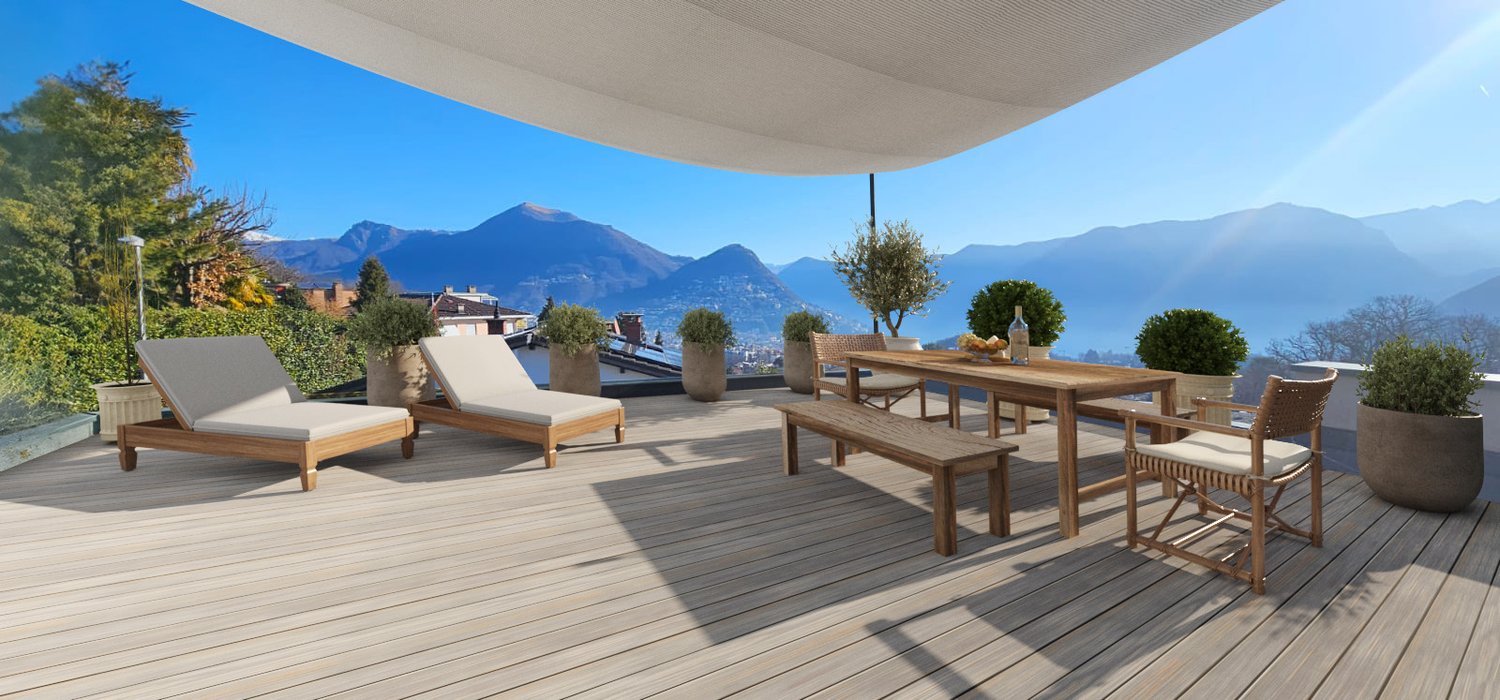 Large wooden deck with outdoor furniture, including lounge chairs and a dining table, with a panoramic view of the mountains and lake in the background. The deck is covered by a canopy and surrounded by potted plants and trees.