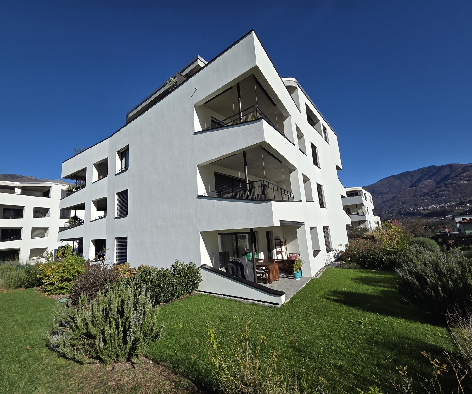 Modern apartment building, 4 stories, white exterior, balconies, gardens in front.
