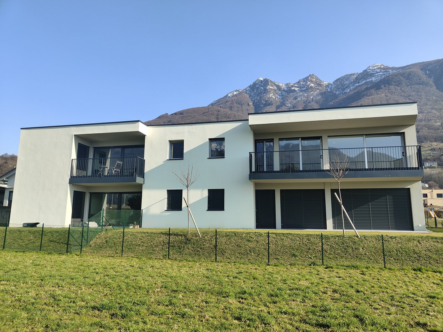 Two-story apartment building with a modern, minimalist design. The building has large windows, balconies, and a garage. It is surrounded by a grassy lawn and has a scenic mountain backdrop.