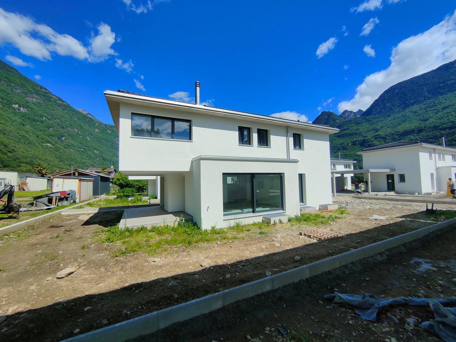 Two-story modern house with large windows, surrounded by mountains and greenery. The house has a simple, clean design with a flat roof and a chimney. There is an open yard or garden area in front of the house.