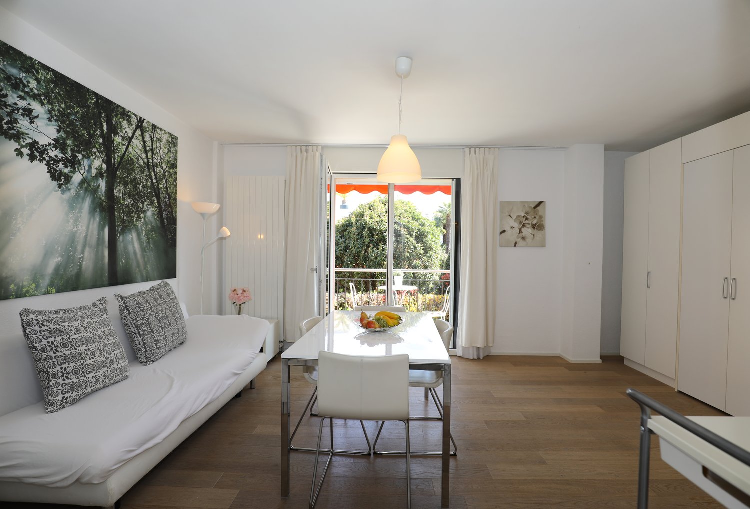 modern living room with white couch, dining table, white chairs, white walls, wooden floor, balcony, white curtains, white lamp, picture on wall