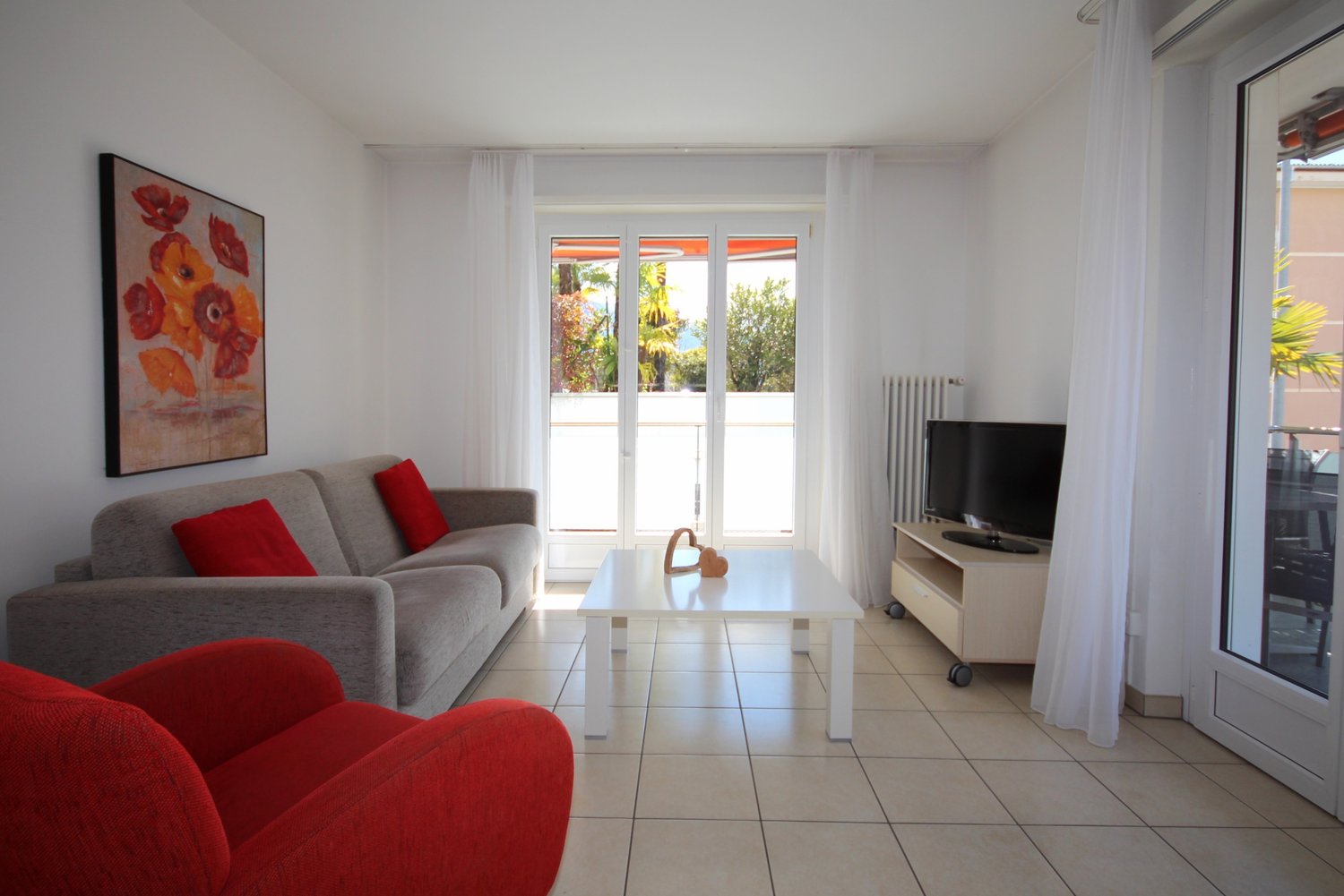 modern living room, gray sofa, red armchair, white coffee table, TV, white curtains, balcony, view of trees