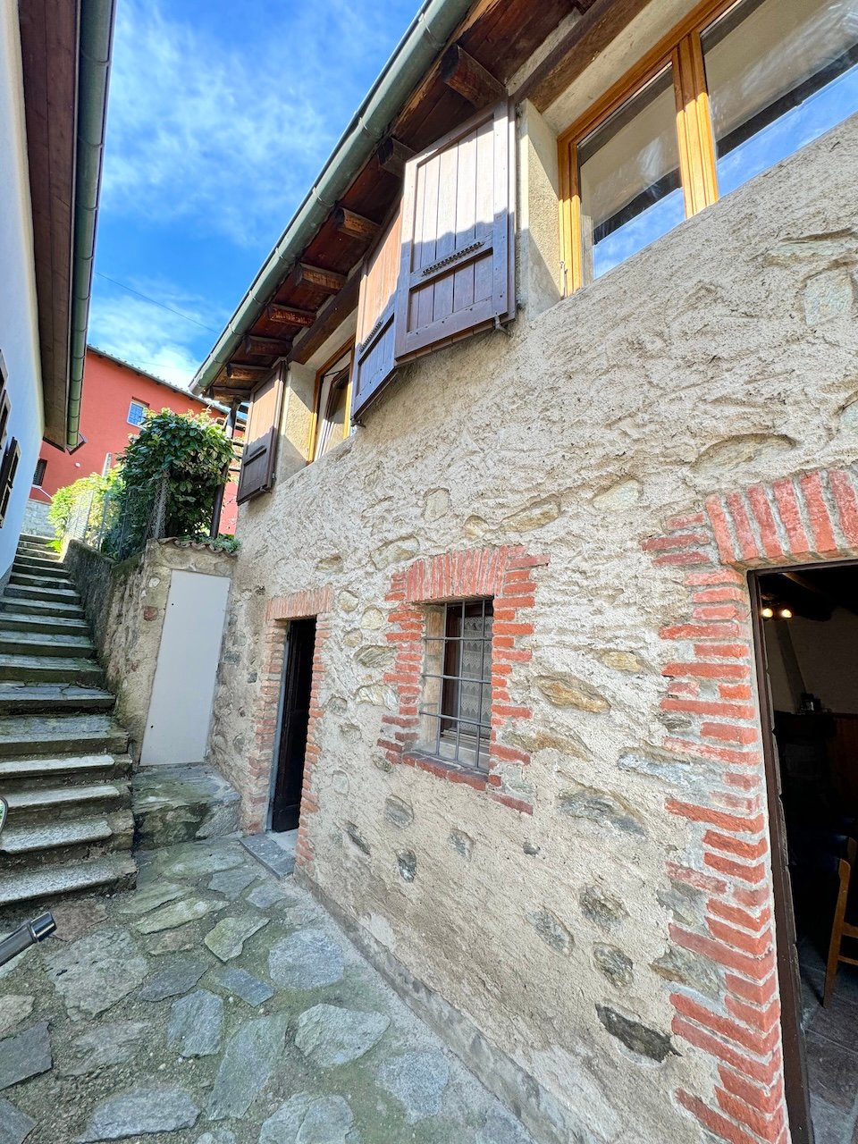 The image shows the exterior of an old stone building with wooden beams and shuttered windows. The building has a stone facade with some brick accents, and there is a set of stairs leading up to the entrance. The surrounding area appears to be a quaint, h