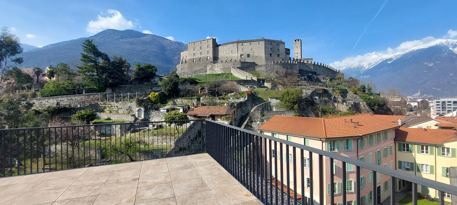 Outdoor area with tiled floor, railings, historical castle in the background, mountains, greenery