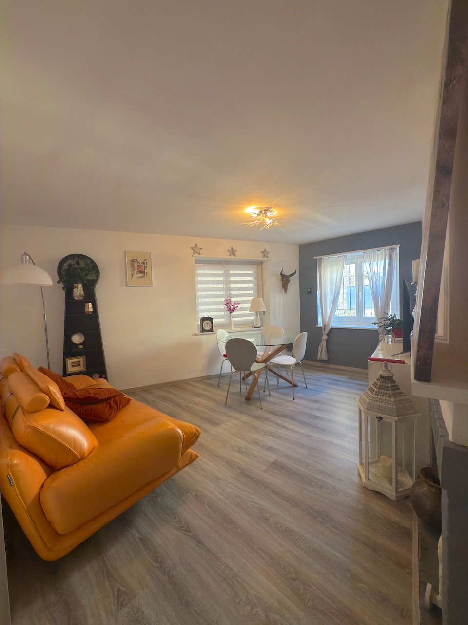 orange couch, dining table with white chairs, wooden floor, white walls, ceiling lamp, potted plant, window with curtains