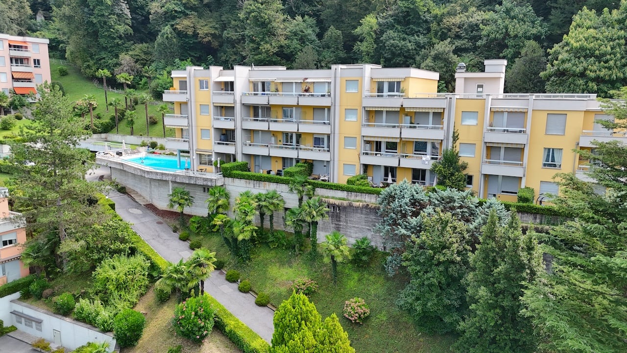 A large yellow apartment building with several balconies. Visible swimming pool. Trees, bushes, and greenery surround the building. In the distance a hilly background is visible.