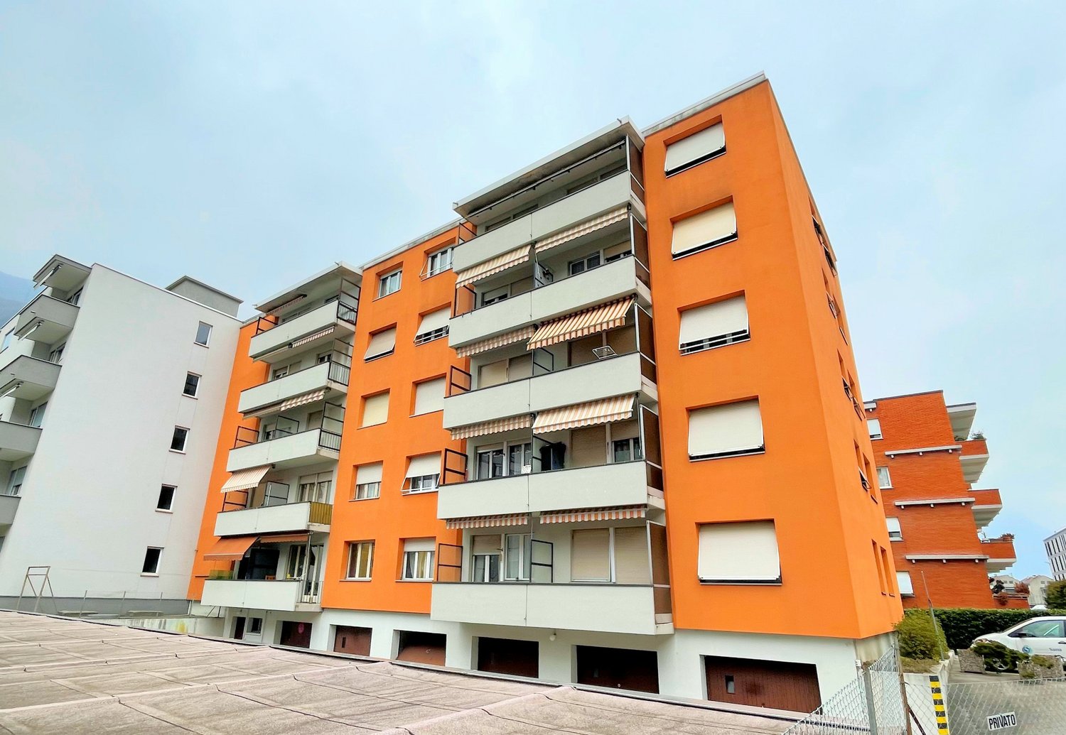 Multi-story apartment building with orange and white exterior, balconies, and parking spaces on the ground level.