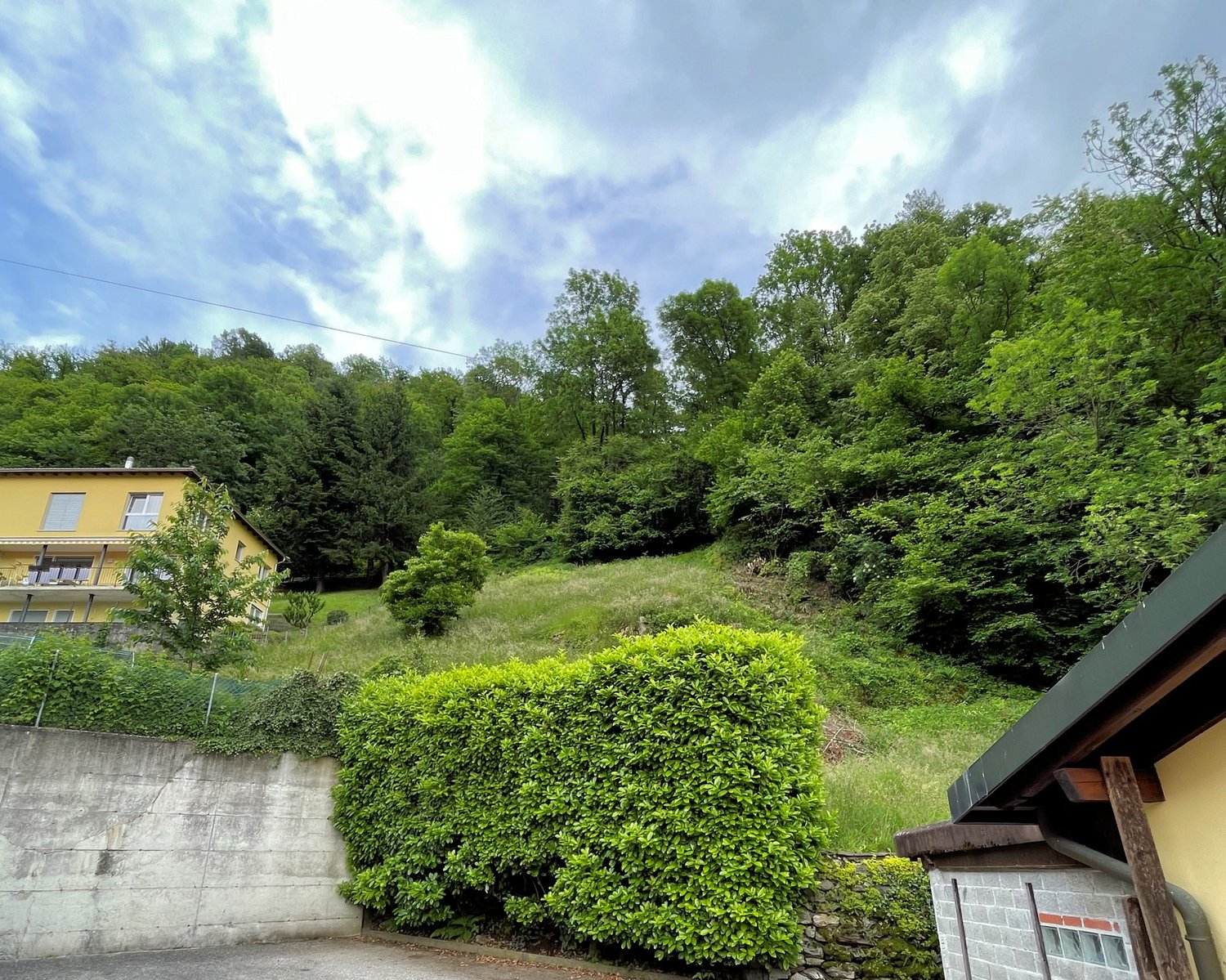 Yellow house with balcony, concrete wall, hedge, green hill, trees
