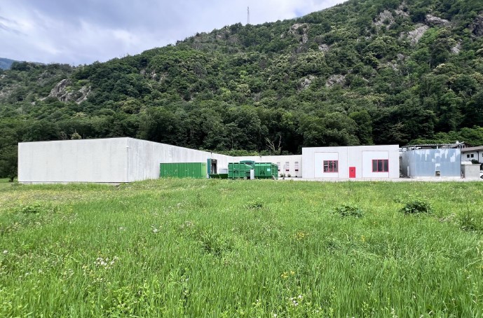 white building, green containers, red doors, large grassy field, mountain in the background