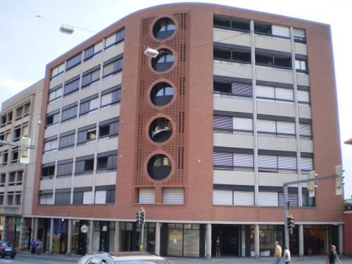 A tall multistory building with a brick facade, large arched windows, and three circular patterns on the front, with people walking on the sidewalk in front