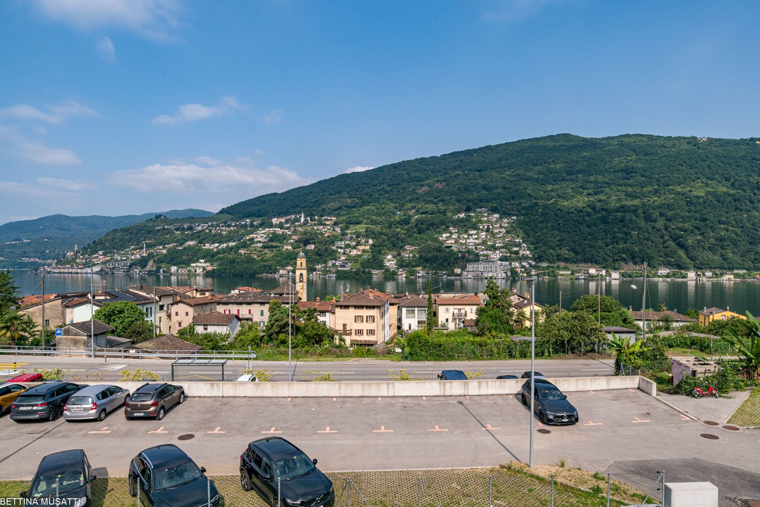 The image shows a scenic view of a lakeside town surrounded by lush, green hills. The town features colorful buildings, a church steeple, and a harbor with boats. There is a parking lot in the foreground with several cars parked. The overall scene depicts