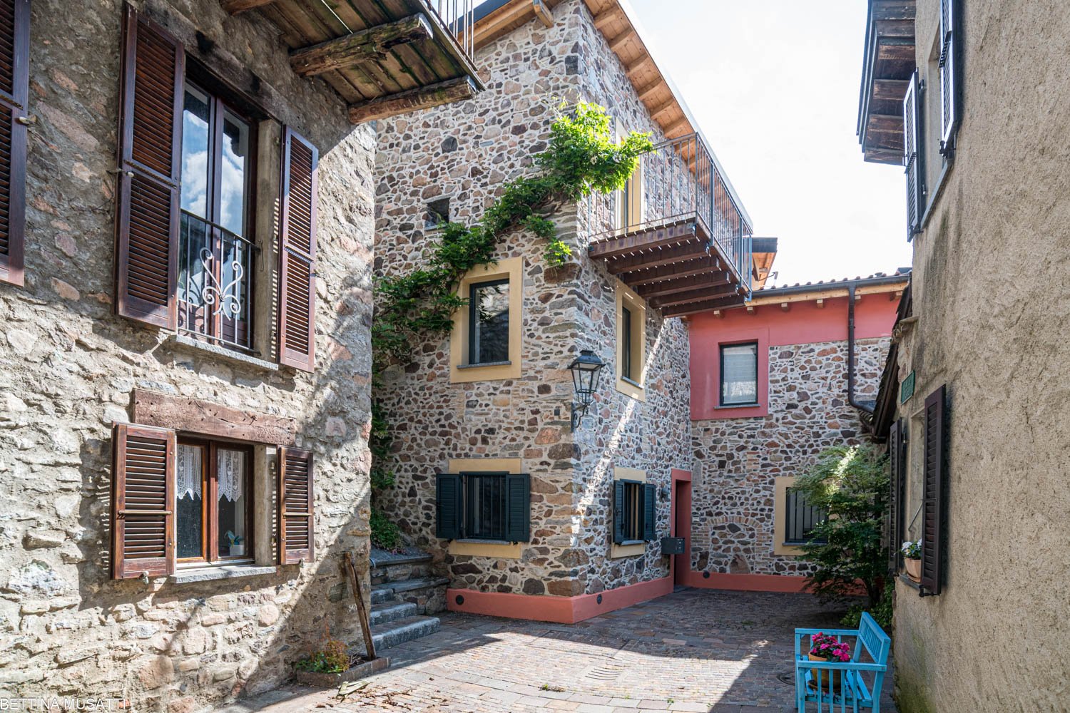 This image shows a traditional stone building with wooden shutters and balconies. The building has a rustic, Mediterranean-style architecture with a cobblestone pathway leading to the entrance. There are potted plants and a small bench visible, adding to