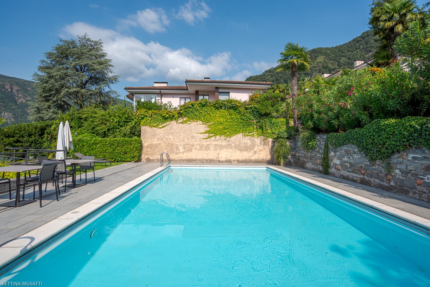 Two-story house with a swimming pool, surrounded by lush greenery and palm trees. The house has a balcony or terrace overlooking the pool area.