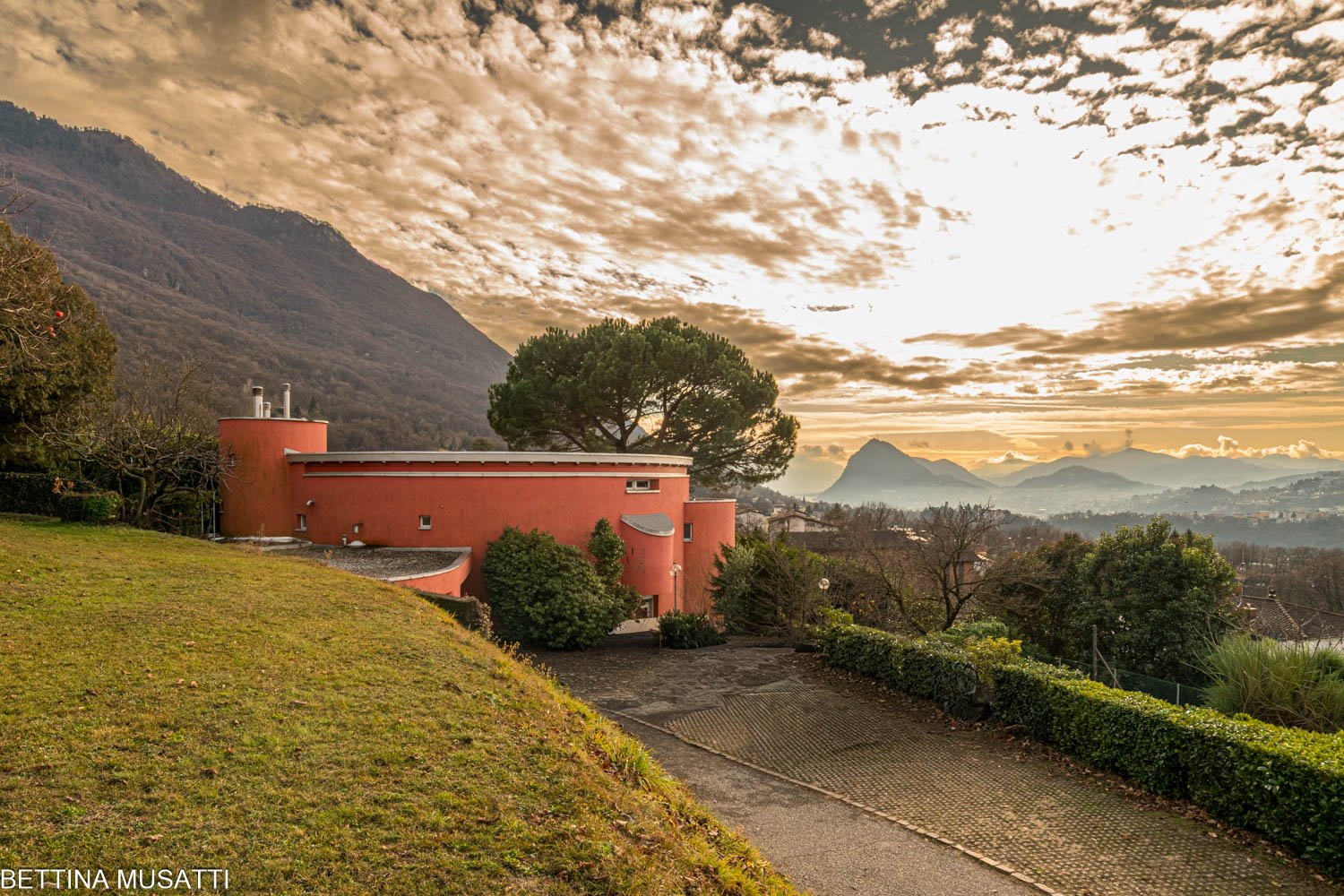 red circular house, circular driveway, mountain backdrop, sunset sky