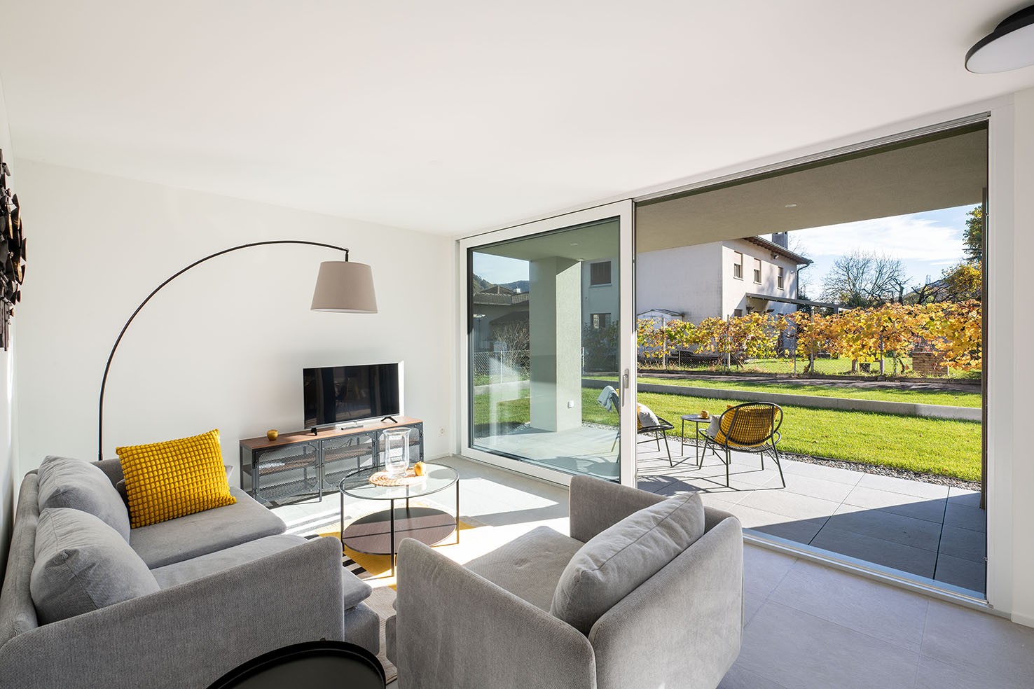 modern living room with sliding glass doors, large windows, gray couches, glass table, yellow pillows, TV, floor lamp, balcony with chairs and table