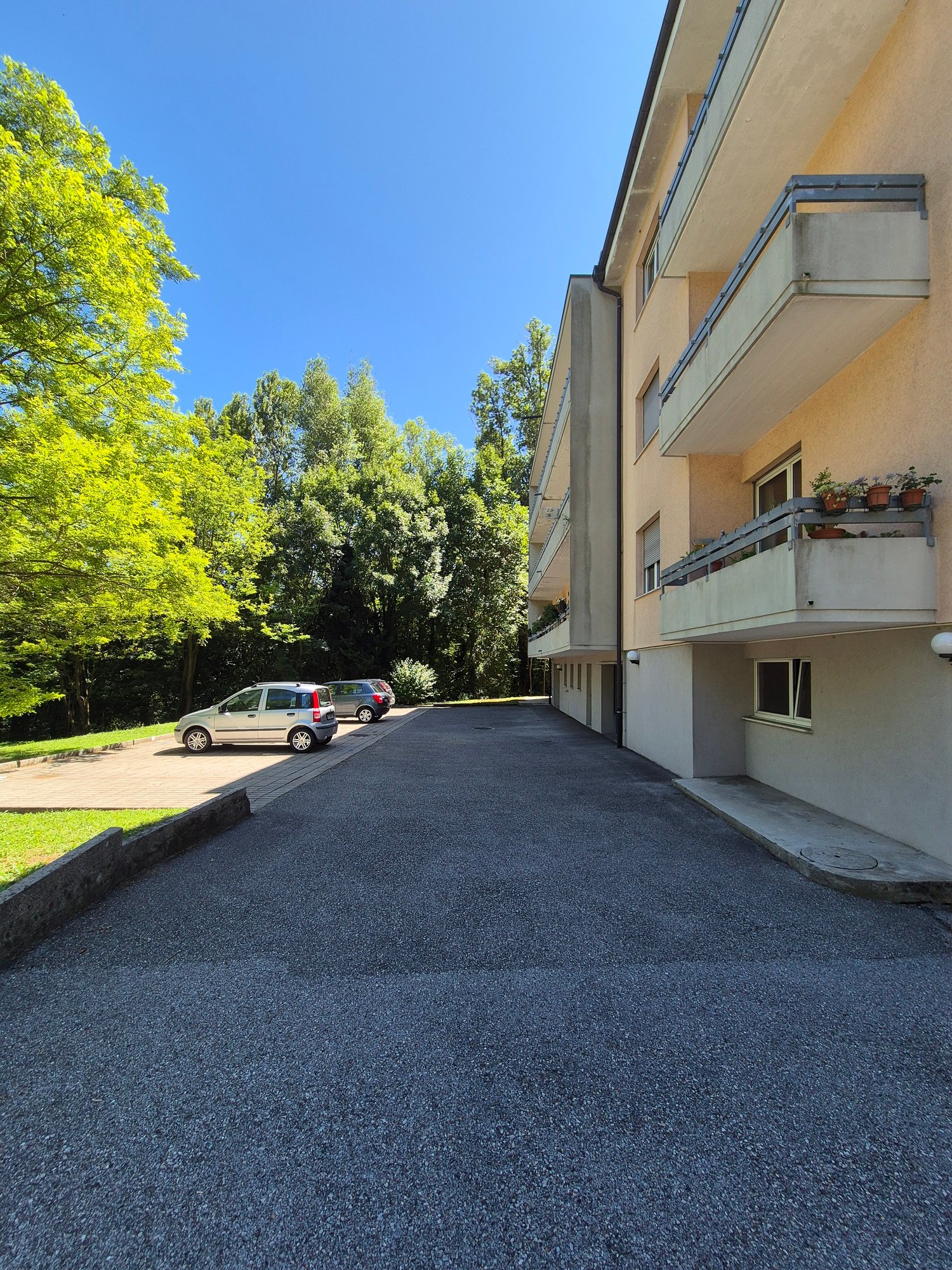 Multi-story apartment building with balconies, surrounded by trees and greenery, with a paved parking area in the foreground.