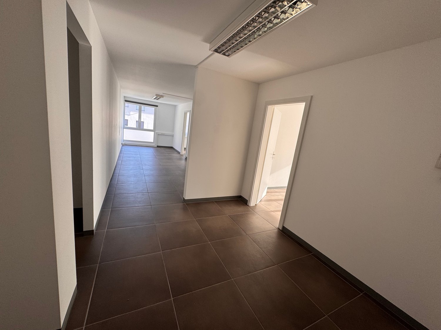 Empty hallway with brown tiled flooring, white walls, windows, and ceiling lighting