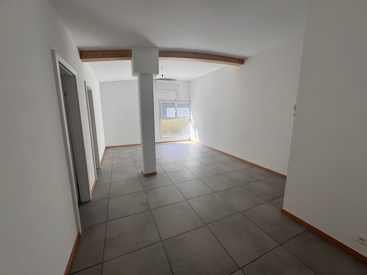 Empty room with white walls, gray tiled floor, wooden beam on ceiling, window with blinds, and an air conditioning unit.