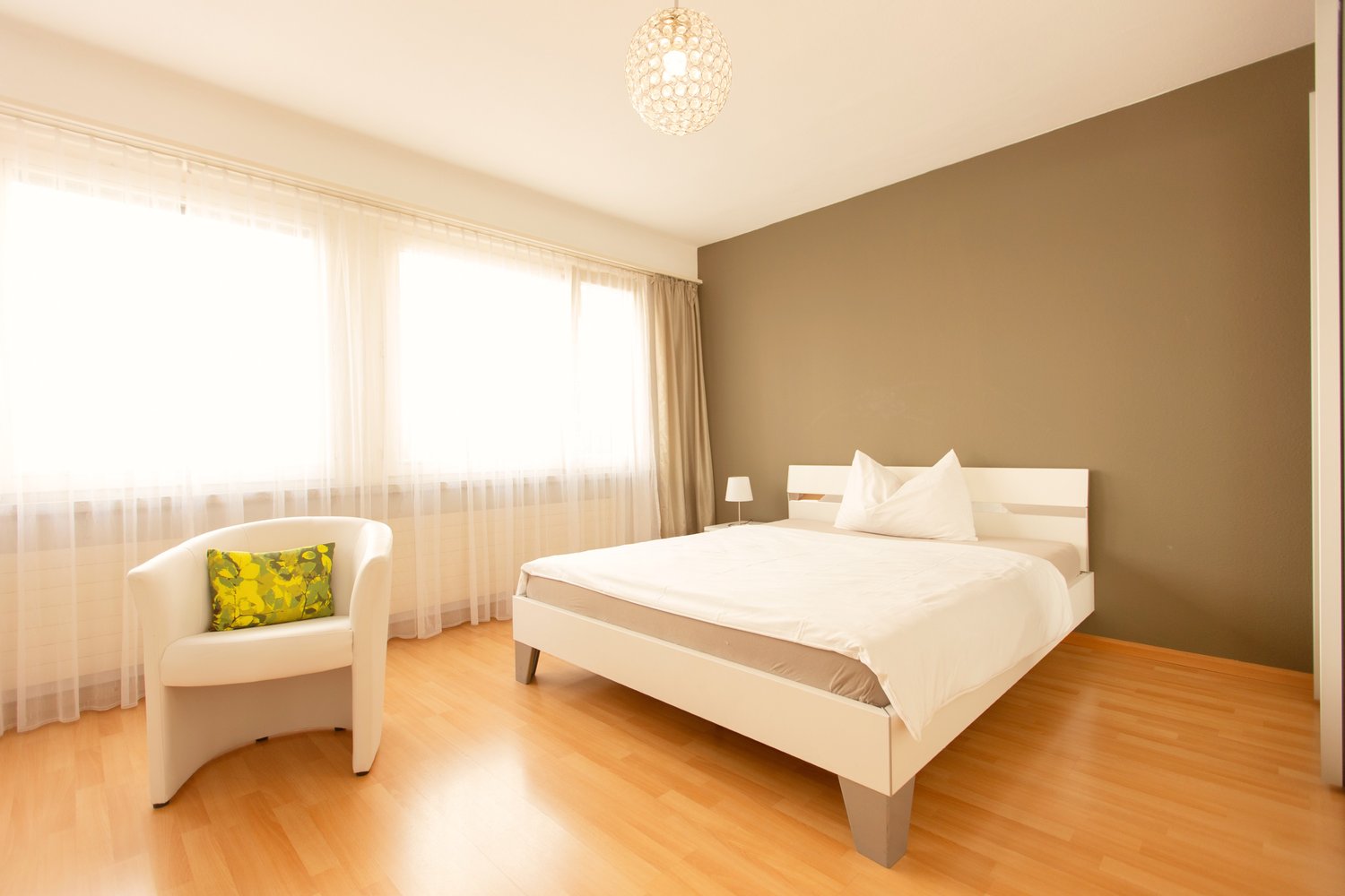 Spacious bedroom with hardwood flooring, beige walls, large windows with sheer curtains, a king-size bed, a white armchair, and a crystal chandelier overhead.
