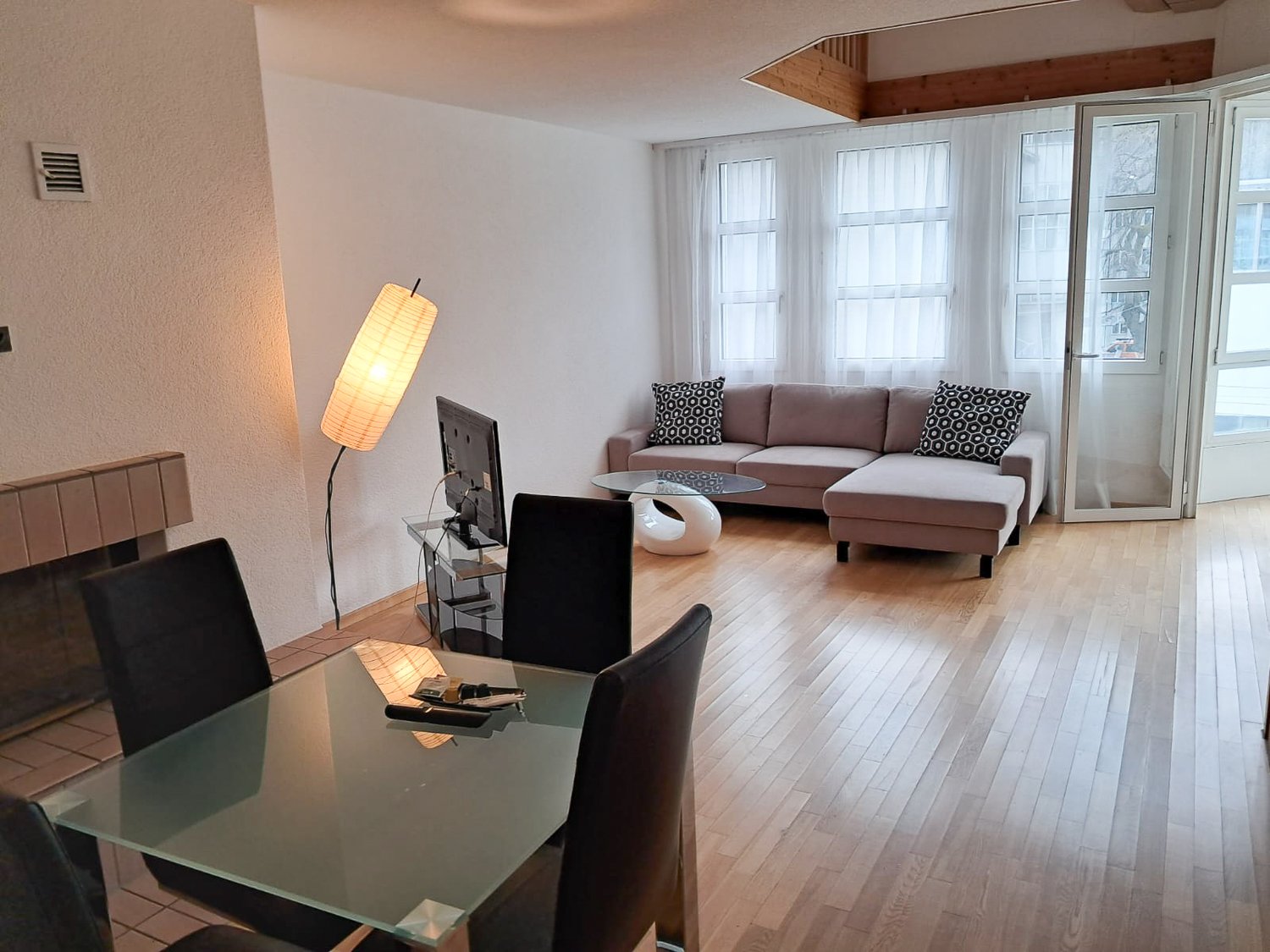 Spacious living room with large windows, hardwood floors, a brown sectional sofa with patterned throw pillows, a glass coffee table, and a floor lamp. The room has an open and airy feel.