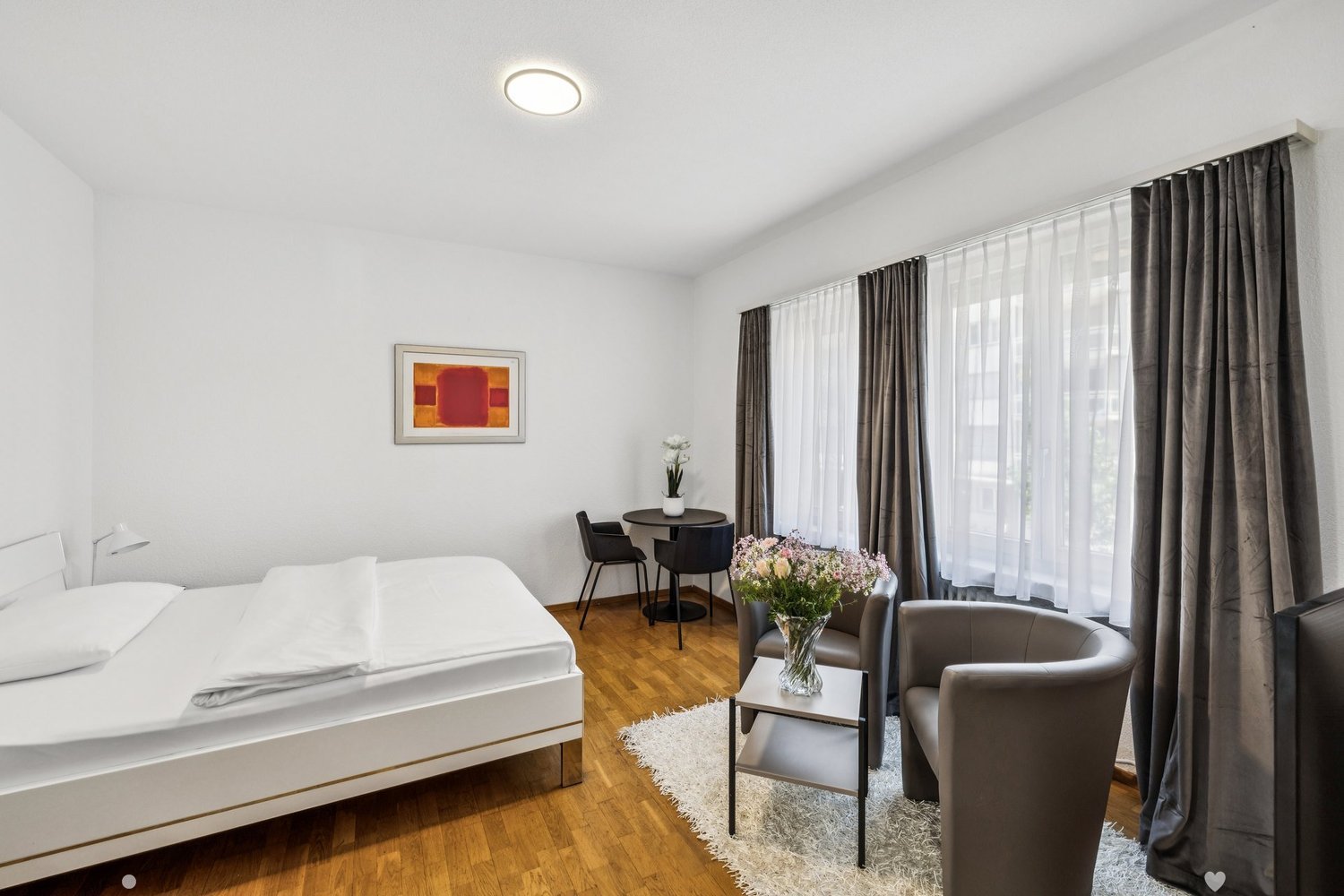 Spacious bedroom with hardwood flooring, large windows with curtains, a double bed, a small round table with two chairs, and a vase of flowers on a side table.