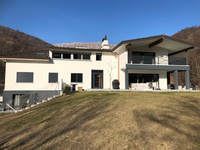 Modern two-story white house on a hill, flat roof, double-story balcony with railings, large lawn in front, trees and mountains in the background.