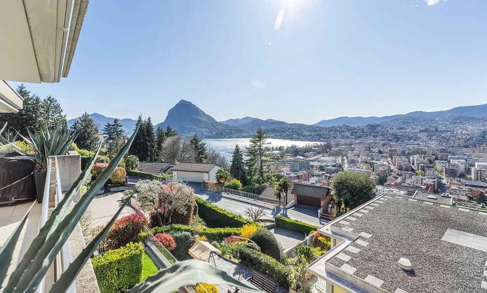 Aerial view of a house with a balcony, surrounded by a garden with various plants and trees, overlooking a cityscape with mountains in the distance.