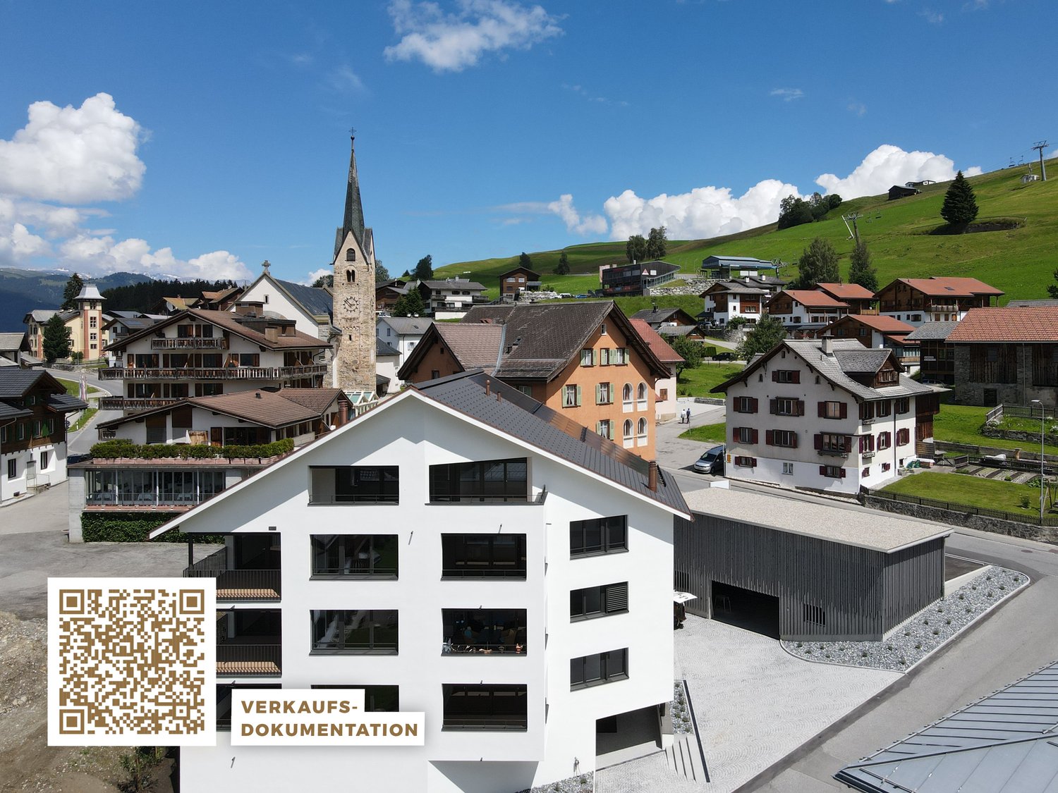 A modern multi-story building with a large parking area in the foreground. The building has a sleek, contemporary design with large windows. In the background, there is a traditional alpine village with a church steeple and other historic buildings nestle