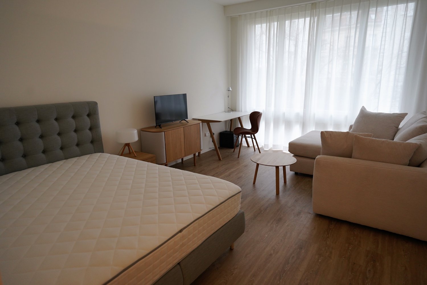A bedroom with a bed, a desk, a chair, and a TV. There is a couch on the right side of the room and a side table next to it. The room has a wooden floor and white walls.
