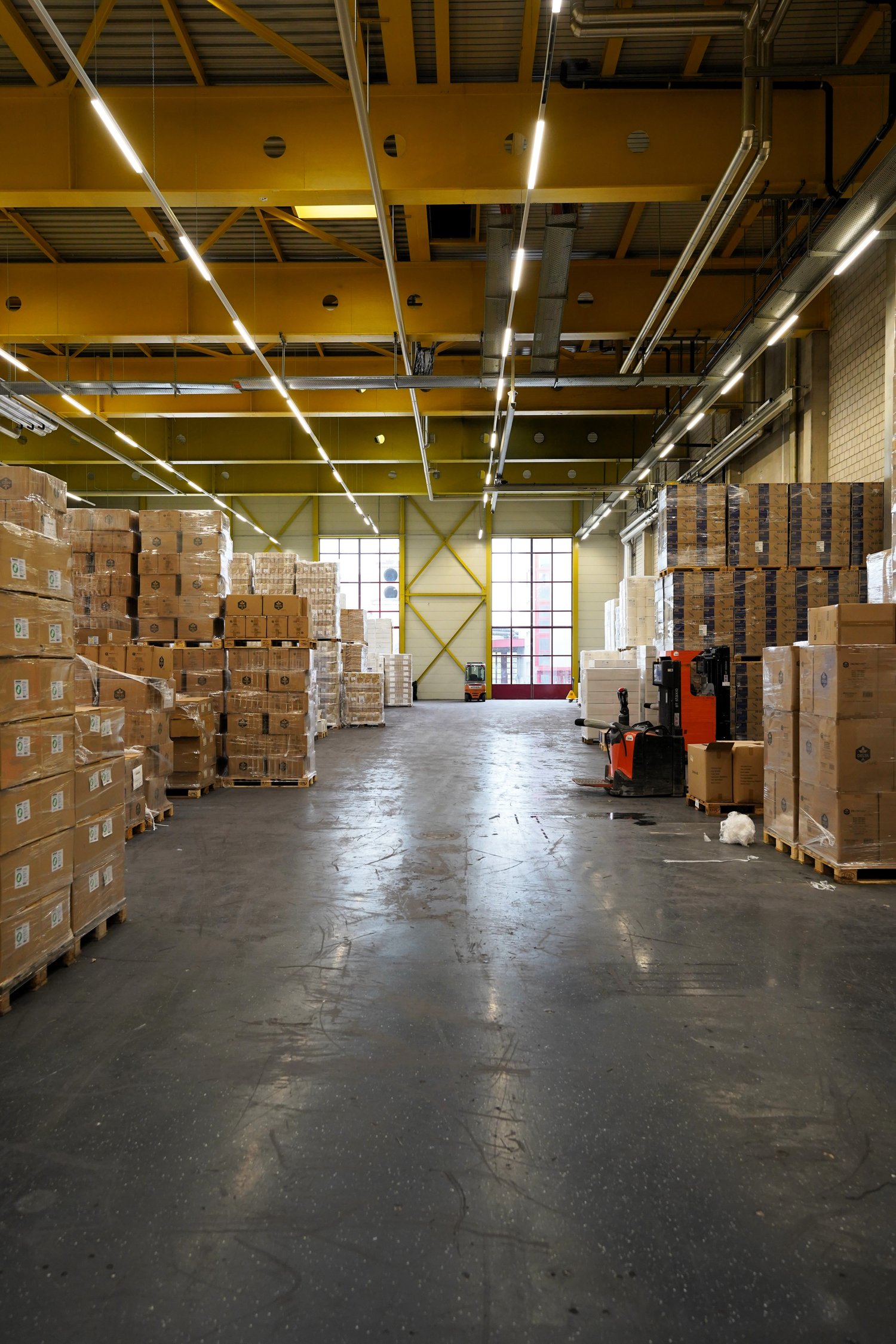 warehouse, large open space, metal roof, large stacks of cardboard boxes, forklifts, windows, bright fluorescent lights