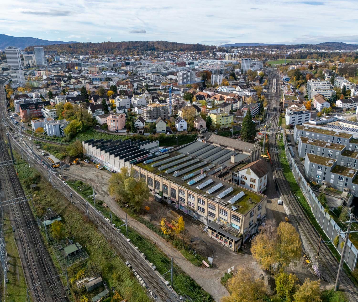 Large factory building with solar panels on the roof, surrounded by a fence, train tracks, and several buildings in the background.