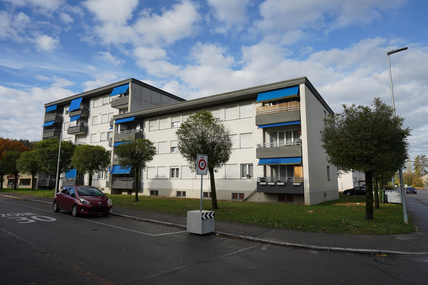 residential building, white exterior, several balconies, red car parked nearby, 30 km/h sign