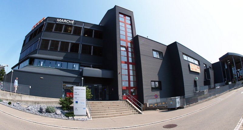 Three-story office building, black exterior, red accents, large windows, stairs leading to entrance, plants and rocks outside.