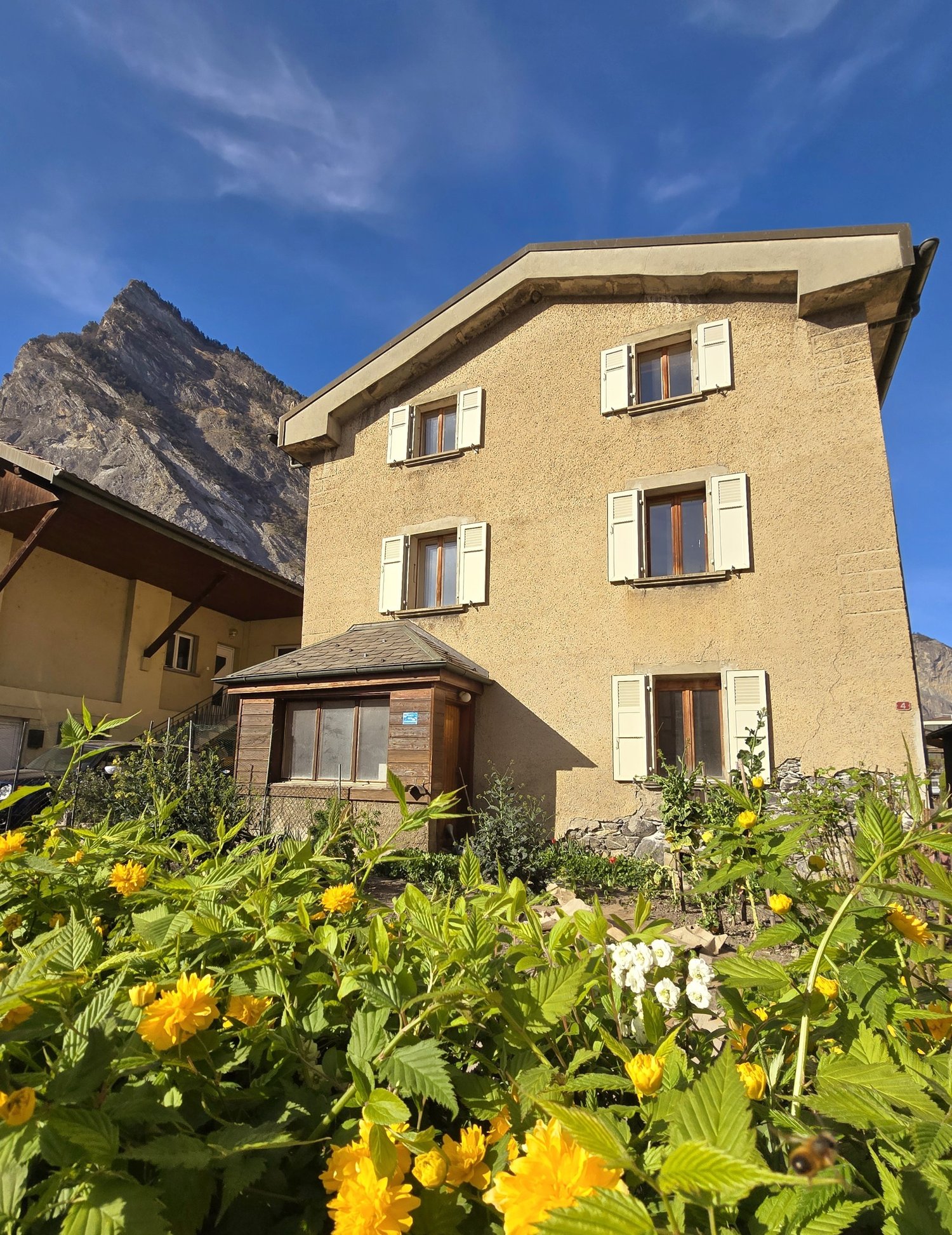 two-story stone house, white shutters, large flower beds with yellow flowers, stunning mountain views